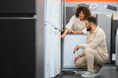 Bir çift, elektronik mağazasında buzdolabını dikkatle inceliyor ve evleri için satın alma kararları veriyor.