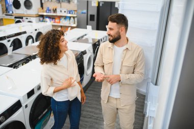 Elektronik mağazasından buzdolabı seçen mutlu çift ev aletleri alırken tartışıyor ve gülümsüyor.