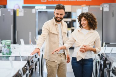 Bir çamaşır makinesini işaret eden mutlu çift, büyük bir elektronik mağazasında ev aletlerini seçiyor.