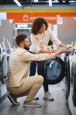 Çift bir elektronik mağazasında çamaşır makinelerini inceliyor, ev aletlerinin alımı için seçenekleri ve özellikleri tartışıyor