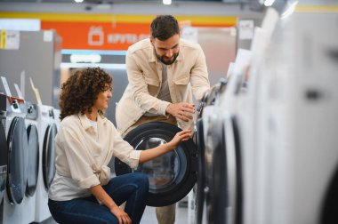 Genç çift, ev aletleri mağazasında yeni çamaşır makinesi seçiyor ve ev eşyaları için önemli bir karar veriyorlar.