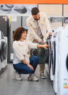 Çift elektronik mağazasında çamaşır makinelerini inceliyor, özellikleri tartışıyor ve ev aletleri satın alma konusunda kararlar alıyor