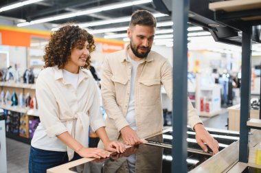 Mutlu çift bir elektronik mağazasından modern mutfak aletleri alırken, bir indüksiyon fırını seçiyor ve geziniyor.