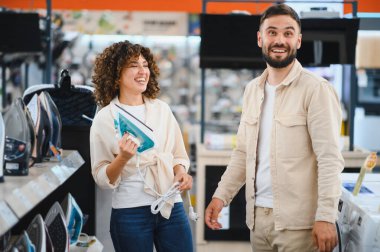 Gülümseyen bir çift elektronik mağazasında ev aletlerini seçiyor, ütüye odaklanıyor. Alışveriş yapmayı ve birlikte karar vermeyi seviyorlar.