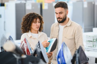 Elektronik mağazasında yeni ütü seçen genç çift, elinde alet tutan kadın, ürün özellikleri hakkında dükkan asistanı açıklamasını dinleyen erkek.