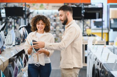 Genç bir çift elektronik mağazasında yeni bir ütü seçiyor. Kadın elinde yeni bir alet tutarken satış asistanı ona tavsiyede bulunuyor.