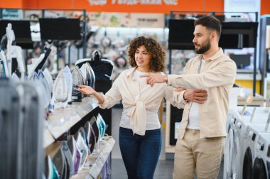 Genç bir çift elektronik mağazasında yeni ütü seçiyor, bir kadın erkeklere aletleri gösteriyor, ev aletleri için alışveriş yapıyor.