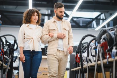 Genç bir çift, parlak bir elektronik mağazasında kol kola yürüyor. Evleri için çeşitli elektrikli süpürgeleri araştırıyor ve karşılaştırıyorlar.
