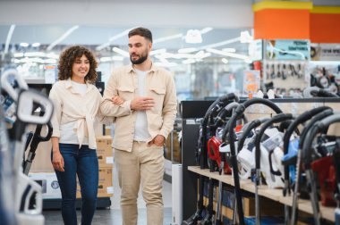 Genç çift ev aletleri mağazasında geziyor ve elektrik süpürgesi seçiyor. Birlikte alışveriş deneyimlerinin tadını çıkarıyorlar.