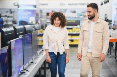 Genç çift bir elektronik mağazasında el ele yürüyor, ev eğlencesi deneyimlerini geliştirmek için en iyi televizyonu arıyorlar.