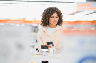 Bir elektronik mağazasında akıllı telefonları karşılaştırırken telefonunu kullanan genç bir kadın satın alma kararı veriyor.