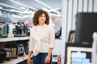 Kadın elektronik mağazasında çeşitli ev aletlerini karıştırıyor. Mutfak ihtiyaçları için en iyi seçenekleri dikkatlice seçiyor.