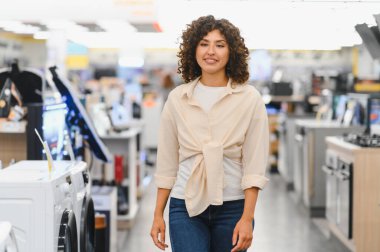 Genç bir kadın elektronik mağazasında yürüyor, ev aletlerini seçiyor, gülümsüyor ve etrafa bakıyor.