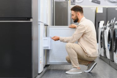 Sakallı adam buzdolabının kapısını açıyor, perakende mağazasından mutfak aletleri alırken dondurucu bölümünü inceliyor.