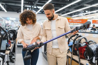 Genç bir çift elektronik mağazasından kablosuz elektrik süpürgesi seçiyor, cihazı inceliyor ve özelliklerini tartışıyor.