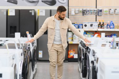 Müşteri, ev aletleri alırken çeşitli çamaşır makinelerini seçenekler dolu kalabalık bir elektronik mağazası koridorunda karşılaştırıyor.