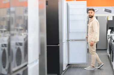 Genç adam elektronik mağazasında buzdolabı kapısını açıyor, çeşitli seçenekler arasından modern ev aletlerini seçiyor.