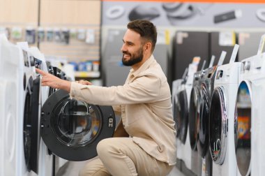 Gülümseyen adam, modern bir elektronik mağazasının alet edevat bölümünde çeşitli çamaşır makinelerini karıştırıyor ve satın alma kararı veriyor.
