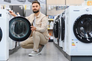 Sakallı bir adam bir mağazada çömelmiş, yeni bir çamaşır makinesini inceliyor ve çamaşır ihtiyaçlarını değerlendiriyor.