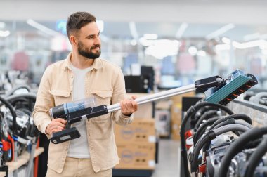 Sakallı adam, perakende mağazasında alışveriş yaparken yeni kablosuz elektrik süpürgesi seçiyor. Seçenekleri değerlendiriyor.