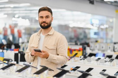 Sakallı adam akıllı telefonları karıştırırken çeşitli model ve markalarla dolu bir elektronik mağazasından yeni bir cep telefonu seçiyor.