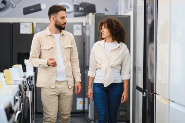 Genç bir çift elektronik mağazasında yürüyor, ev aletlerini seçiyor, alışveriş hakkında tartışıyor ve karar veriyorlar.