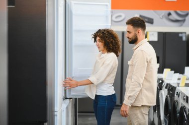 Genç çift ev aletleri mağazasında yeni buzdolabını inceliyor, modern mutfak için satın alma kararı alıyorlar.