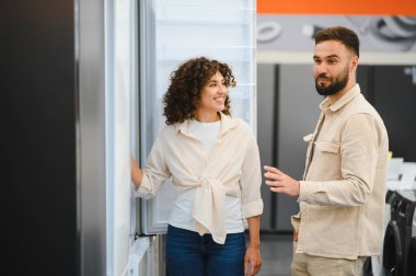 Erkek ve kadın bir elektronik mağazasında ev aletlerini seçerken seçenekleri tartışıyor, buzdolabına odaklanıyor