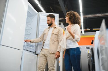 Genç çift eşyalar mağazasında yeni buzdolabı seçiyor, özellikleri tartışıyor ve satın alma kararı alıyorlar.