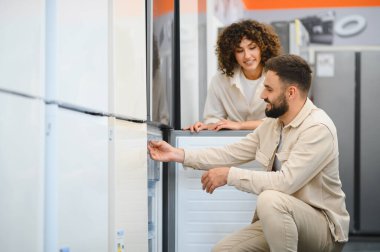 Mutlu çift elektronik mağazasından yeni buzdolabı seçiyor, ev aletlerini inceliyor, gülümsüyor.