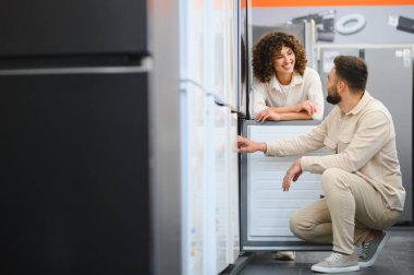 Mutlu çift ev aletleri mağazasında yeni buzdolabı seçiyor, özellikleri tartışıyor ve satın alma kararı alıyorlar.