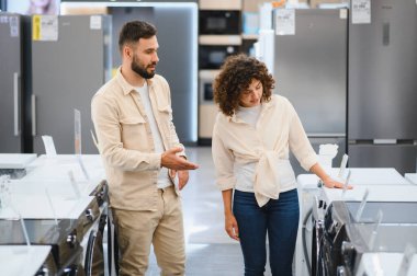 Çift yeni bir çamaşır makinesi seçiyor, konuşuyor ve bir elektronik mağazasında farklı modelleri değerlendiriyor.