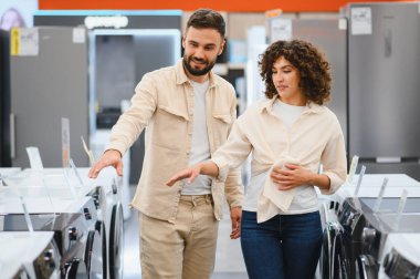 Çift, elektronik mağazasındaki modern ev aletlerini değerlendiriyor ve ev ihtiyaçlarının özelliklerini ve seçeneklerini tartışıyor.