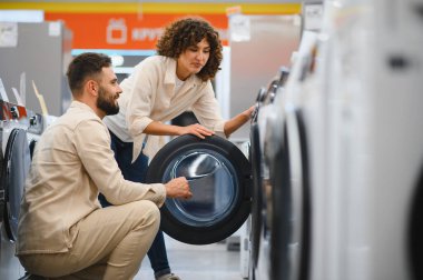 Alet mağazasından yeni çamaşır makinesi seçen genç çift, satın almadan önce ürün özelliklerini inceliyor