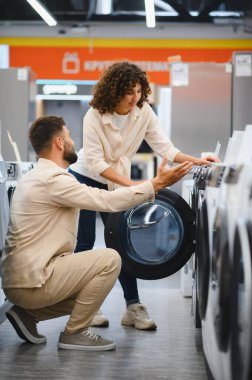 Bir çift elektronik mağazasında çamaşır makinelerini araştırıyor, seçenekleri tartışıyor ve ev aletlerinin alımı hakkında kararlar alıyor.