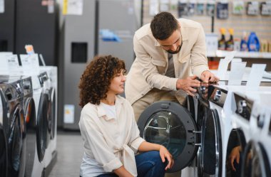 Genç çift yeni çamaşır makinesi seçiyor, özellikleri keşfediyor ve ev aletleri mağazasından alışveriş yapmayı tartışıyorlar.