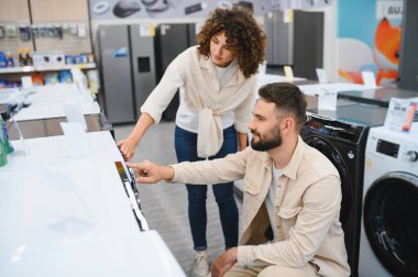 Genç çift, elektronik mağazasında ev aletlerini seçerken çamaşır makinesini işaret ederek alışveriş deneyiminin tadını çıkarıyor.