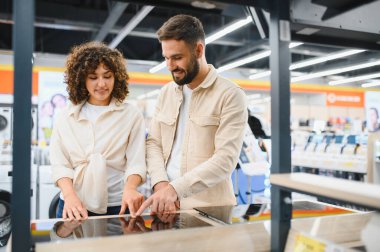 Mutlu çift indüksiyon sobasını işaret ediyor, ev aletleri mağazasından mutfak gereçleri alıyor.
