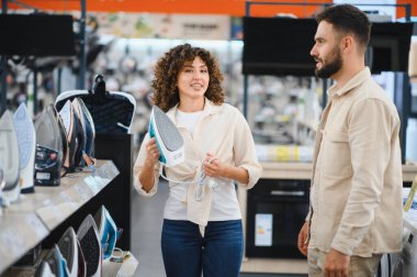 Genç çift, bir elektronik mağazasının ev aletleri bölümünde yeni bir ütü arıyor, seçenekleri tartışıyor ve özellikleri karşılaştırıyor