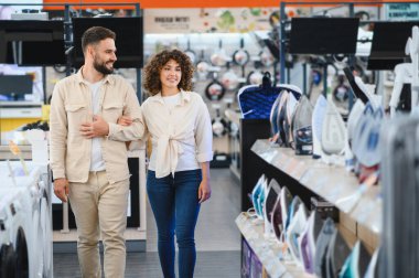 Bir elektronik mağazasında yürüyen mutlu bir çift, ev aletlerini seçerken birlikte eğleniyorlar ve tercihlerini tartışıyorlar.
