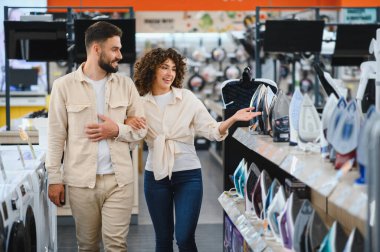 Genç çift, modern bir ev aletleri mağazasının parlak koridorlarına göz atarken yeni bir ütü seçerken mutlu bir şekilde alışveriş deneyimlerinin tadını çıkarıyor.