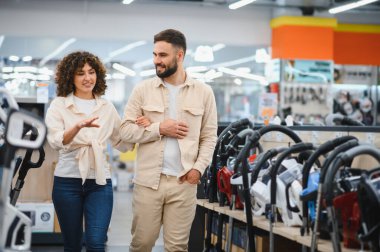 Çift elektronik mağazasında görülüyor, ev aletlerini tartışıyor ve seçiyor, raflarda sergilenen elektrikli süpürgelere odaklanıyor