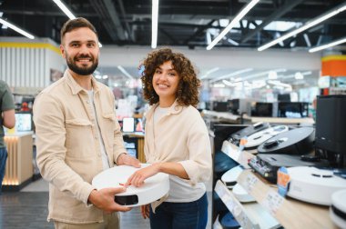Robot elektrikli süpürge tutan mutlu çift elektronik mağazasından ev aletlerini seçiyor.