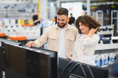 Müşteri elektronik mağazasında alışveriş yaparken büyük ekran televizyonu gösteren mutlu çift.