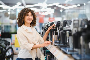 Gülümseyen kadın ev aletleri mağazasında alışveriş yaparken çeşitli kahve makinelerini karıştırıyor. Seçimlerden hoşlanıyor ve seçimler yapıyor.