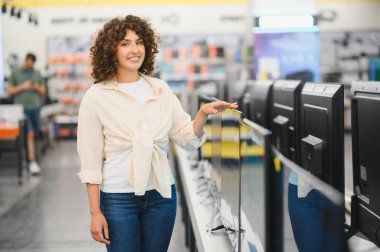 Bir perakende mağazasında elektronik eşya alırken televizyonu işaret eden gülümseyen kadın, farklı modelleri karşılaştırıyor.