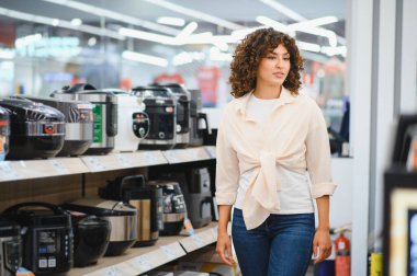 Genç bir kadın elektronik mağazasında yürüyor ve birden fazla fırın seçiyor, raflardaki çeşitli modelleri inceliyor.