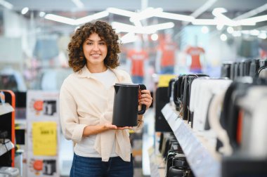 Elektrikli su ısıtıcısı tutan gülümseyen kadın, elektronik mağazasının ev aletleri bölümünde alışveriş yapıyor.
