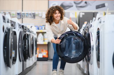 Ev aletleri mağazasında çamaşır makinesini seçerken, satın alma kararı verirken ve ürünü önerirken mutlu müşteri başparmağını kaldırır