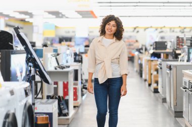 Genç bir kadın elektronik mağazasına giriyor, çeşitli ev aletlerini karıştırıyor ve ev eşyaları için düşünceli seçimler yapıyor.